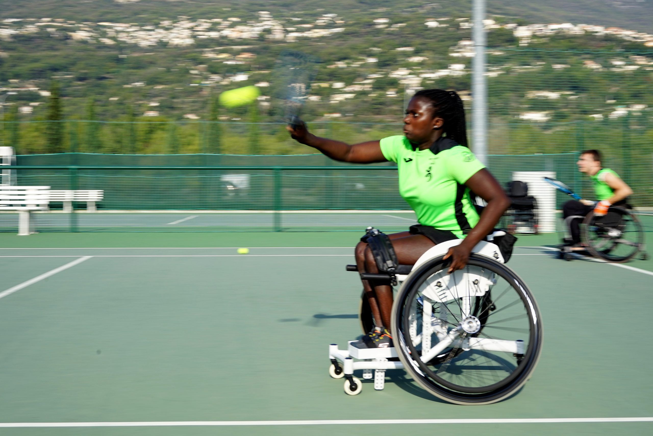 Tennista in carrozzina alza il braccio per colpire la palla su un campo da tennis all’aperto.